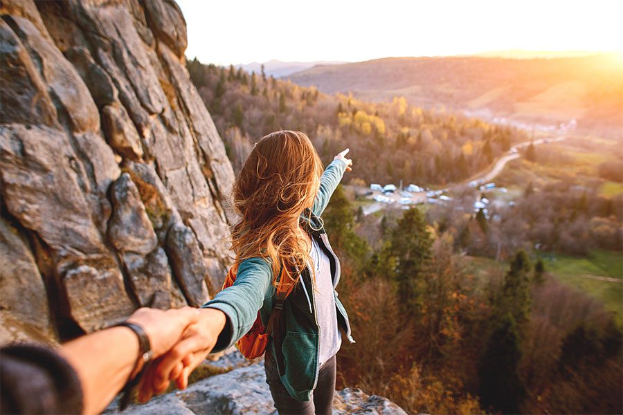 About Our Agency - Scenic View of a Young Woman Holding Hands with Her Partner While Hiking Up a Mountain and Pointing at the Setting Sun Over the Mountains Beyond
