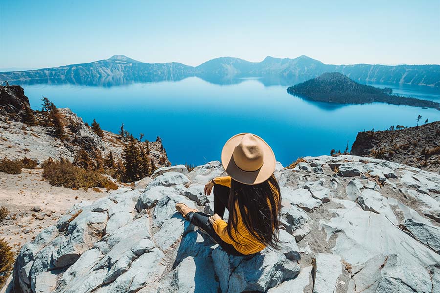 Client Center - View of a Young Woman Sitting on Top of a Mountain While on Vacation Looking Out at the Mountains and Lake