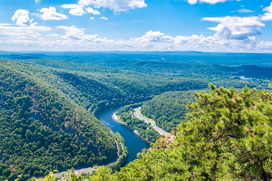 Our Locations - Aerial View of A River Surrounded by Green Mountains Full of Trees Against a Cloudy Blue Sky in Pennsylvania