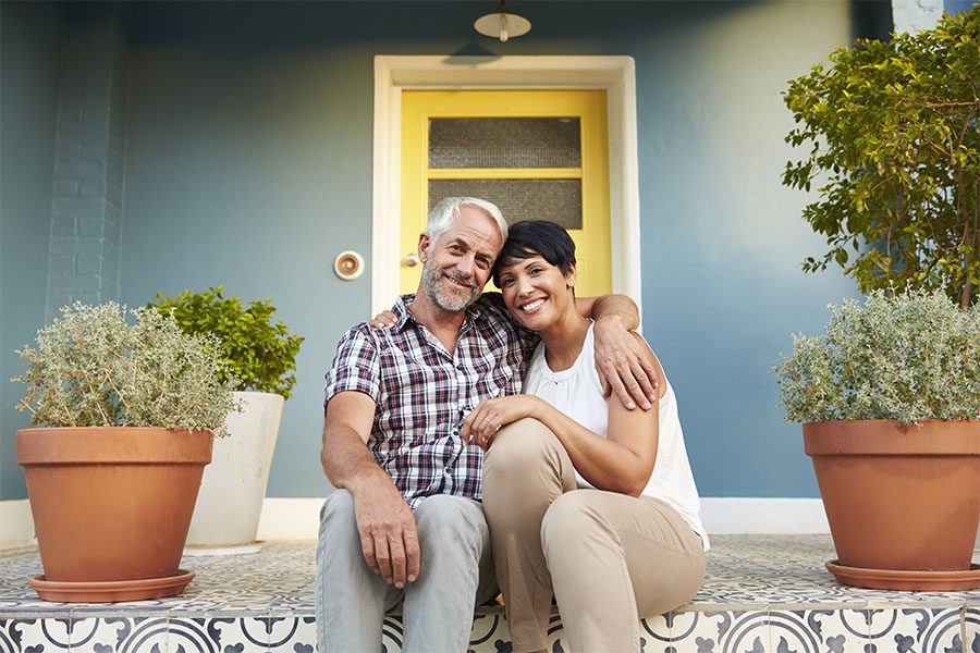 Retirement and Financial Planning - Portrait of a Cheerful Mature Couple Sitting on the Front Steps of Their Home with Multiple Plants Around Them